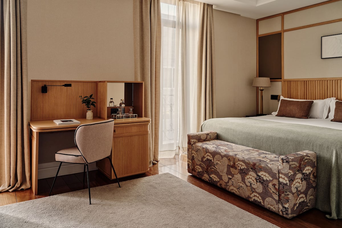 Une chambre d’hôtel avec un bureau, une chaise et un banc en bois, un lit avec des oreillers et une table de chevet avec une lampe.