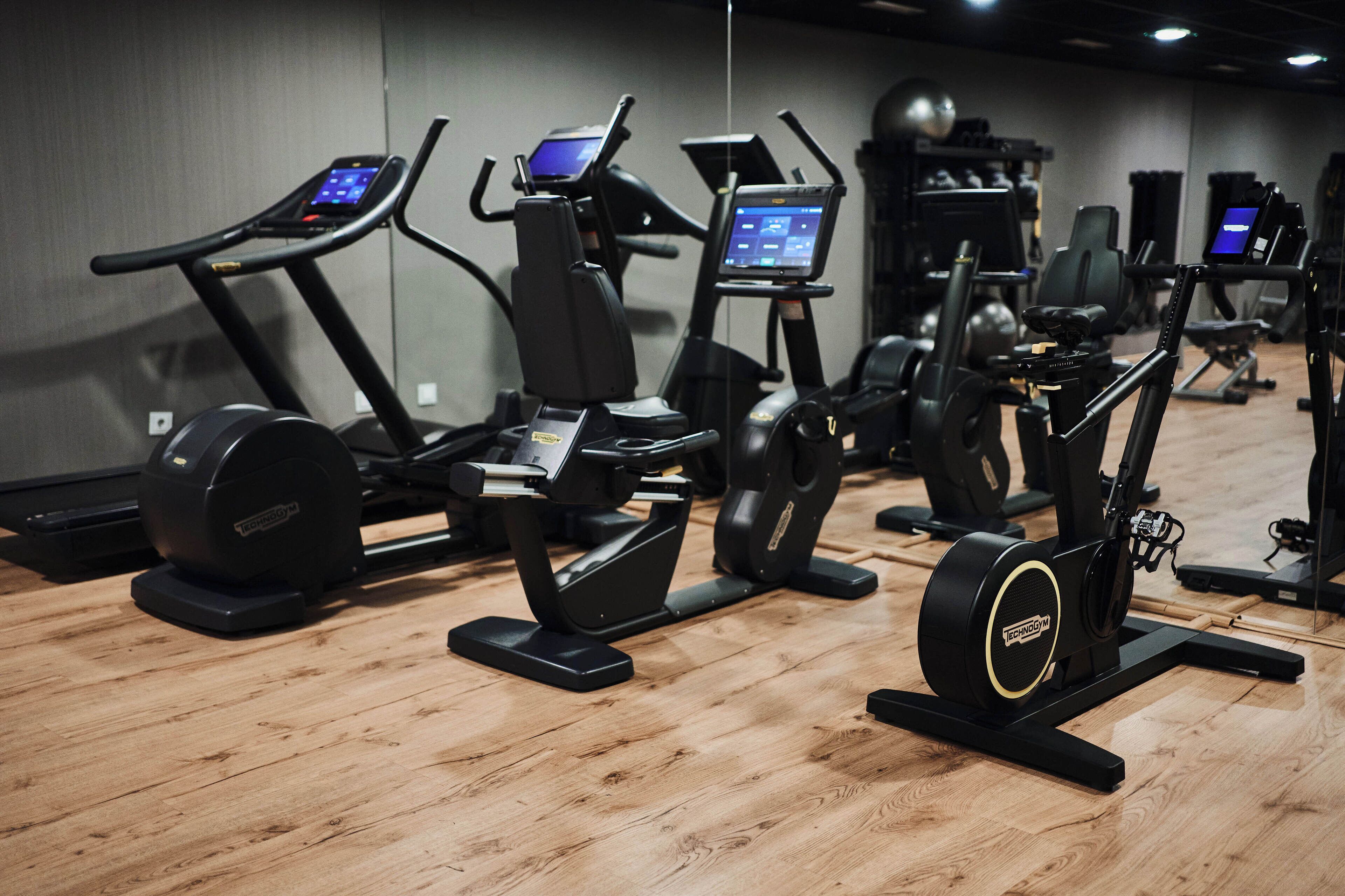 Un gimnasio con diversas máquinas de ejercicio, incluyendo cintas de correr y elípticas, dispuestas en una sala amplia con suelo de madera.