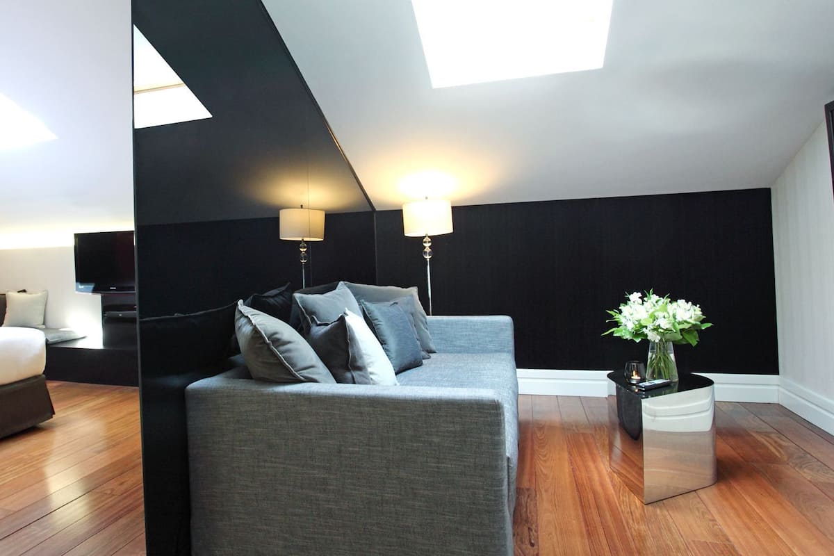 Une chambre d’hôtel moderne avec un lit, un canapé gris, une télévision et une lucarne au plafond.