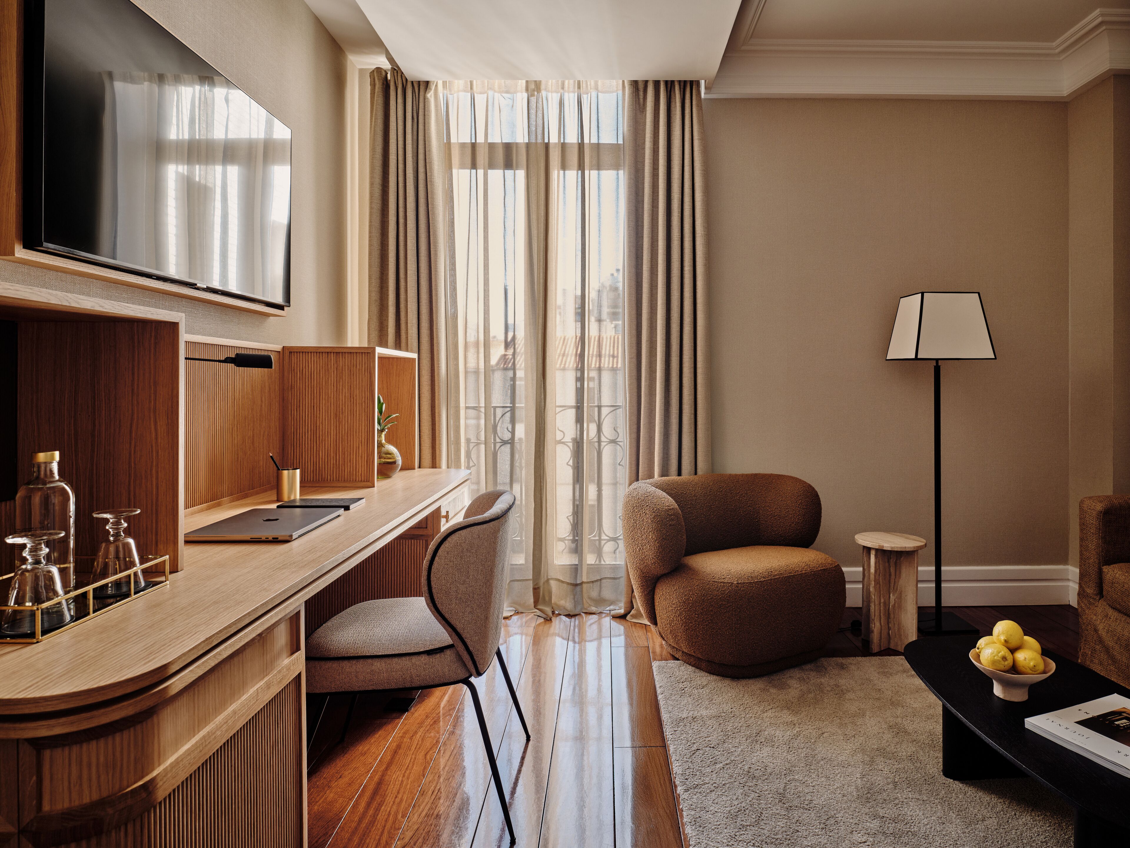 Une chambre d’hôtel moderne avec un bureau en bois, un fauteuil confortable, une table basse ronde ornée de citrons et un lampadaire.