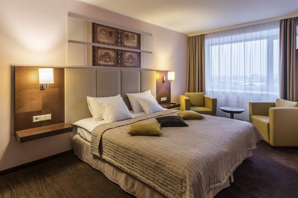 A hotel room with a large bed, two armchairs, a small table, and a window with curtains.