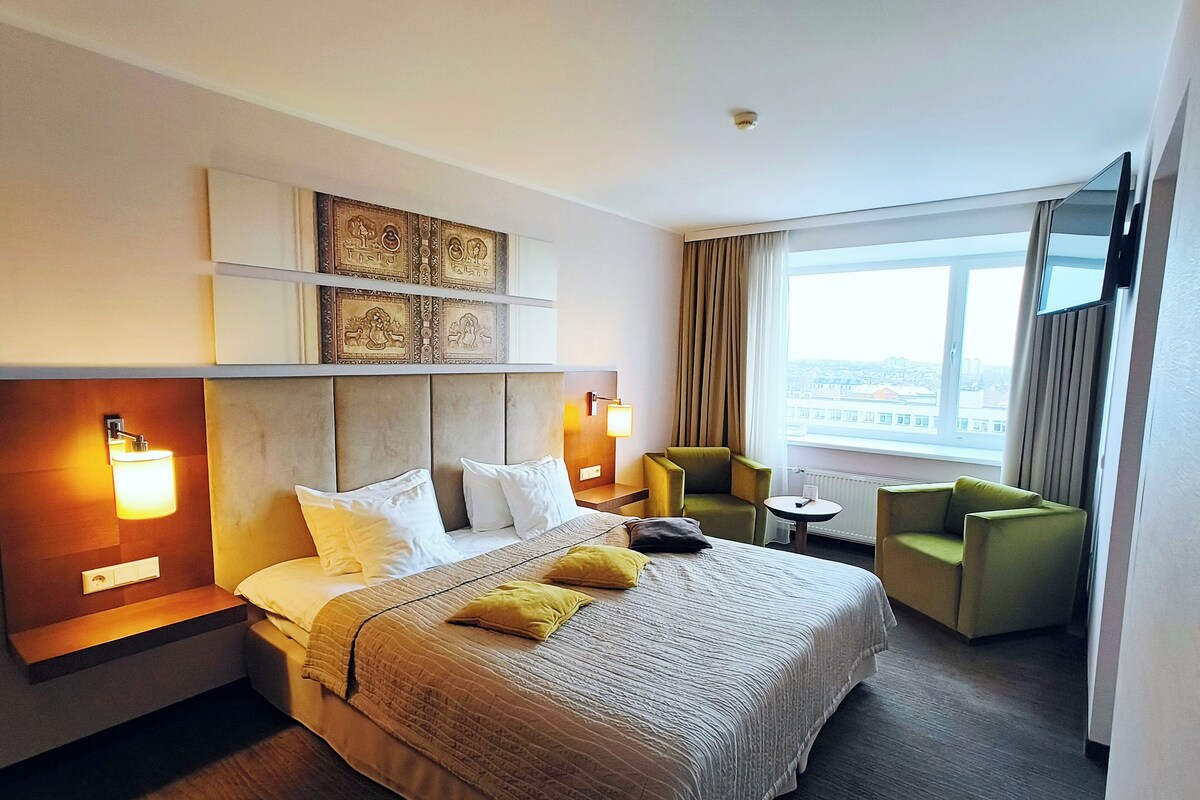 Une chambre d’hôtel avec un grand lit, deux fauteuils, une petite table et une grande fenêtre donnant sur la ville.