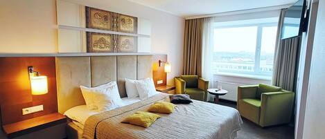 Een hotelkamer met een groot bed, twee fauteuils, een klein tafeltje en een groot raam met uitzicht op de stad.
