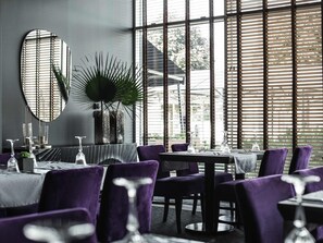 Un espace repas moderne avec des tables rondes, des chaises violettes et de grandes fenêtres équipées de stores.