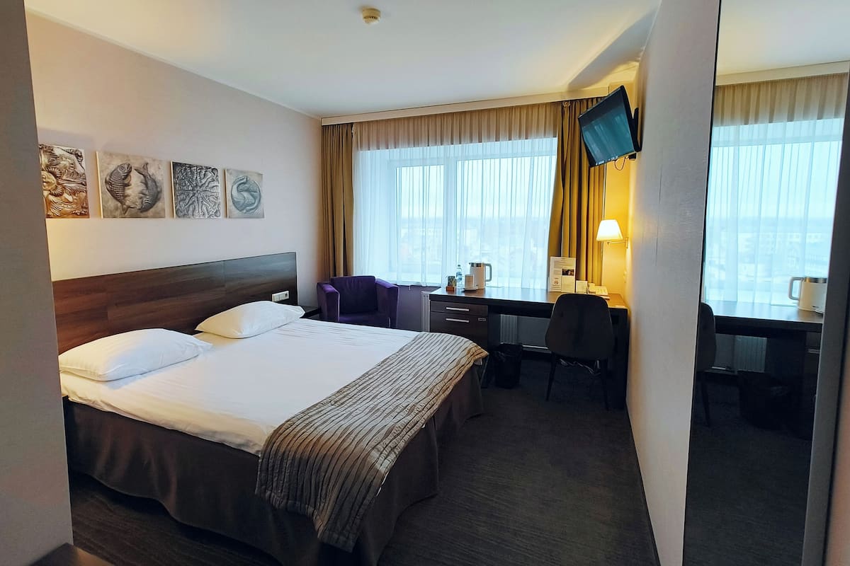 A hotel room with a large bed, a desk with a chair, a TV mounted on the wall, and artwork on the wall.