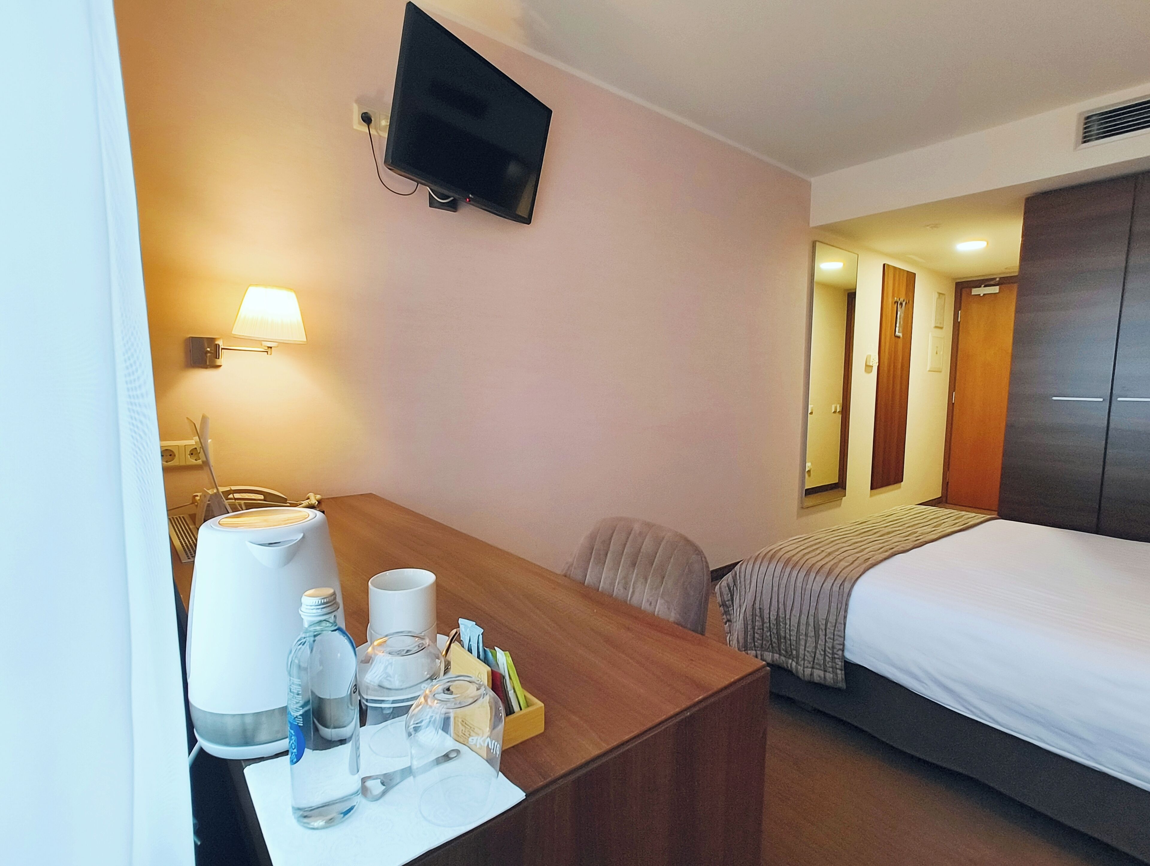 Een hotelkamer met een bed, een bureau met lamp, een aan de muur bevestigde televisie en een kledingkast.