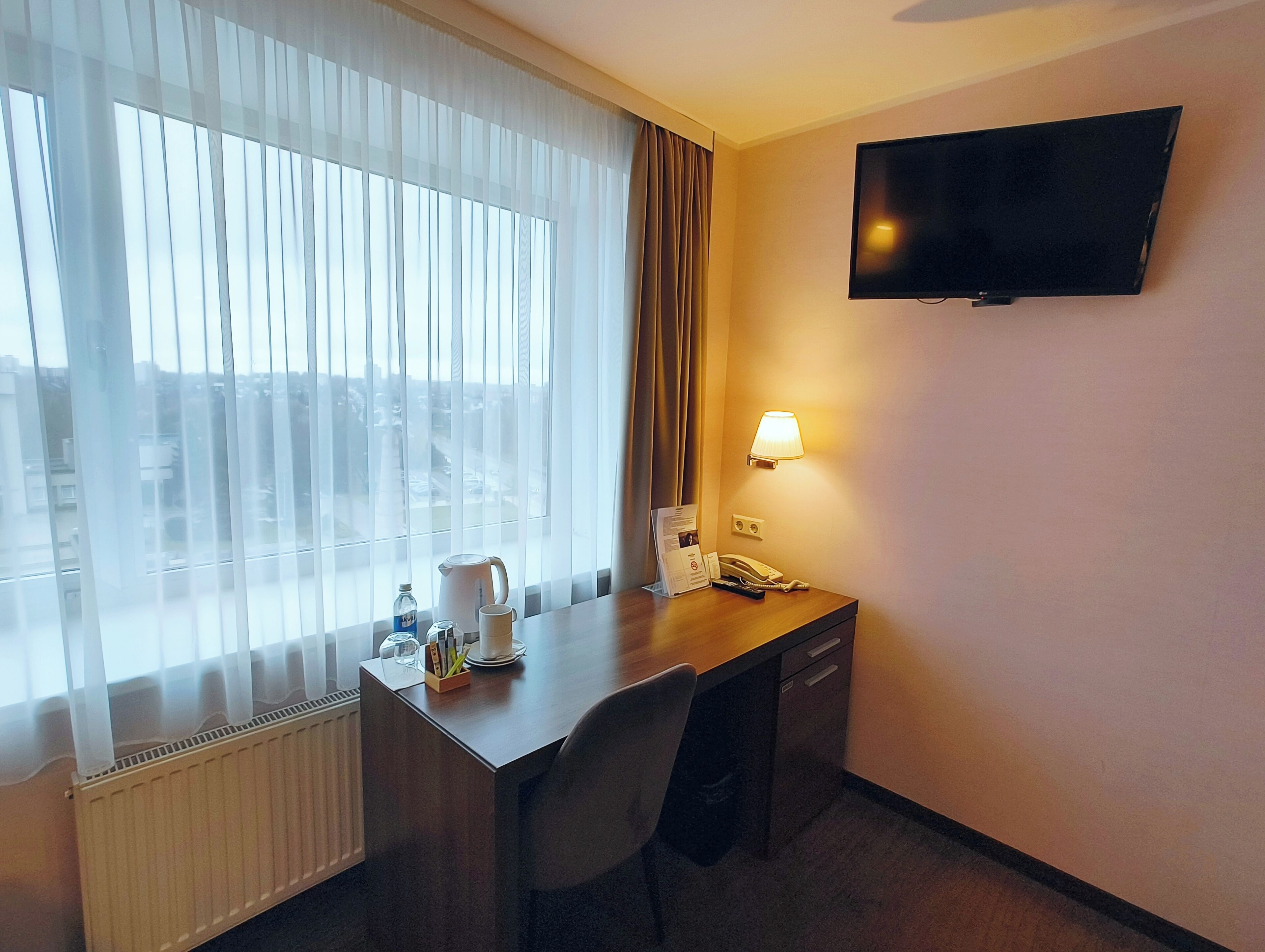 A hotel room with a desk, chair, a flat-screen TV, a lamp, a kettle, a cup, and a bottle of water.