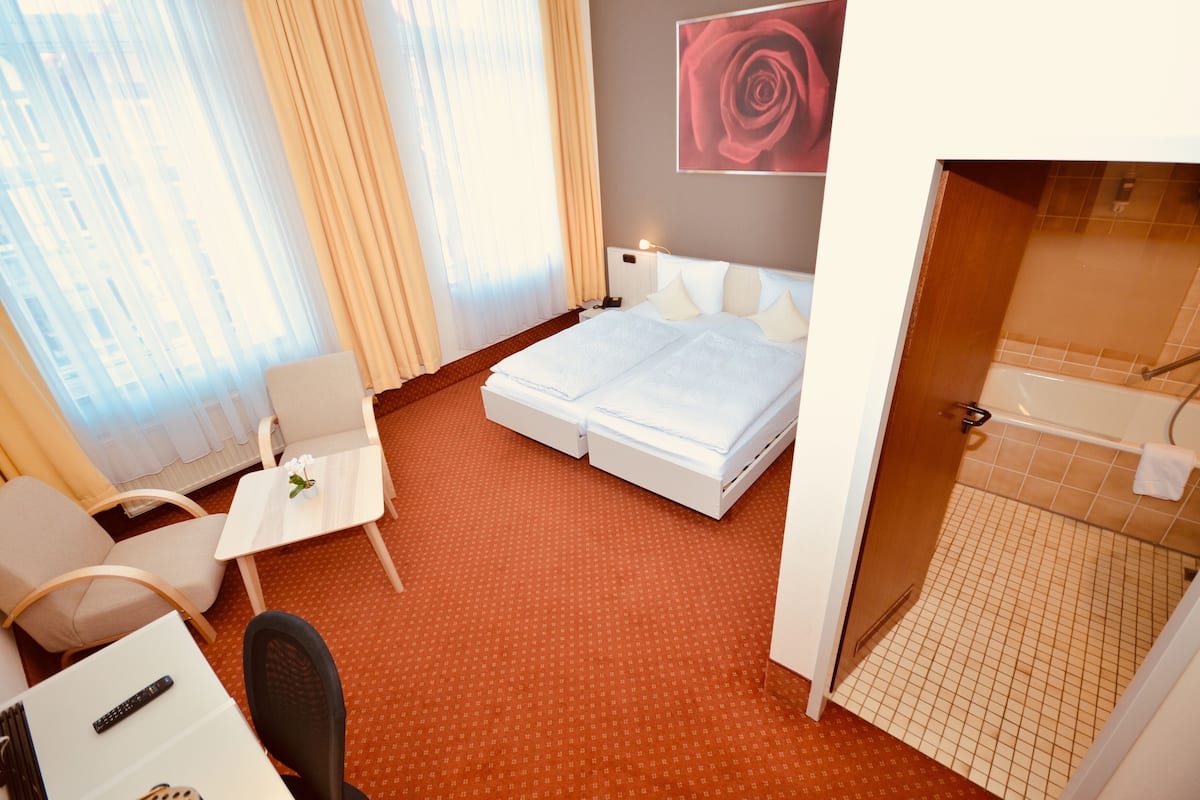Ein Hotelzimmer mit einem Bett, einem kleinen Tisch mit einer Blume, einem Sessel, einem Schreibtisch mit einer Fernbedienung und einer Dusche.