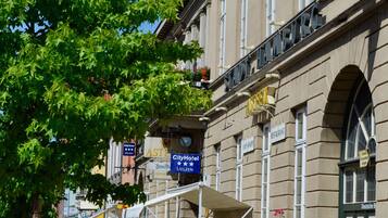 Un hotel con fachada clásica europea, mesas al aire libre y un cielo azul despejado.