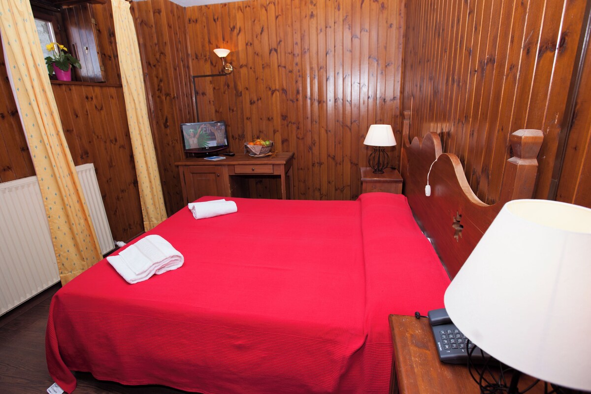 Una camera d'albergo con parete rivestita in legno, un letto con copripiumino rosso, una scrivania con lampada e un televisore.