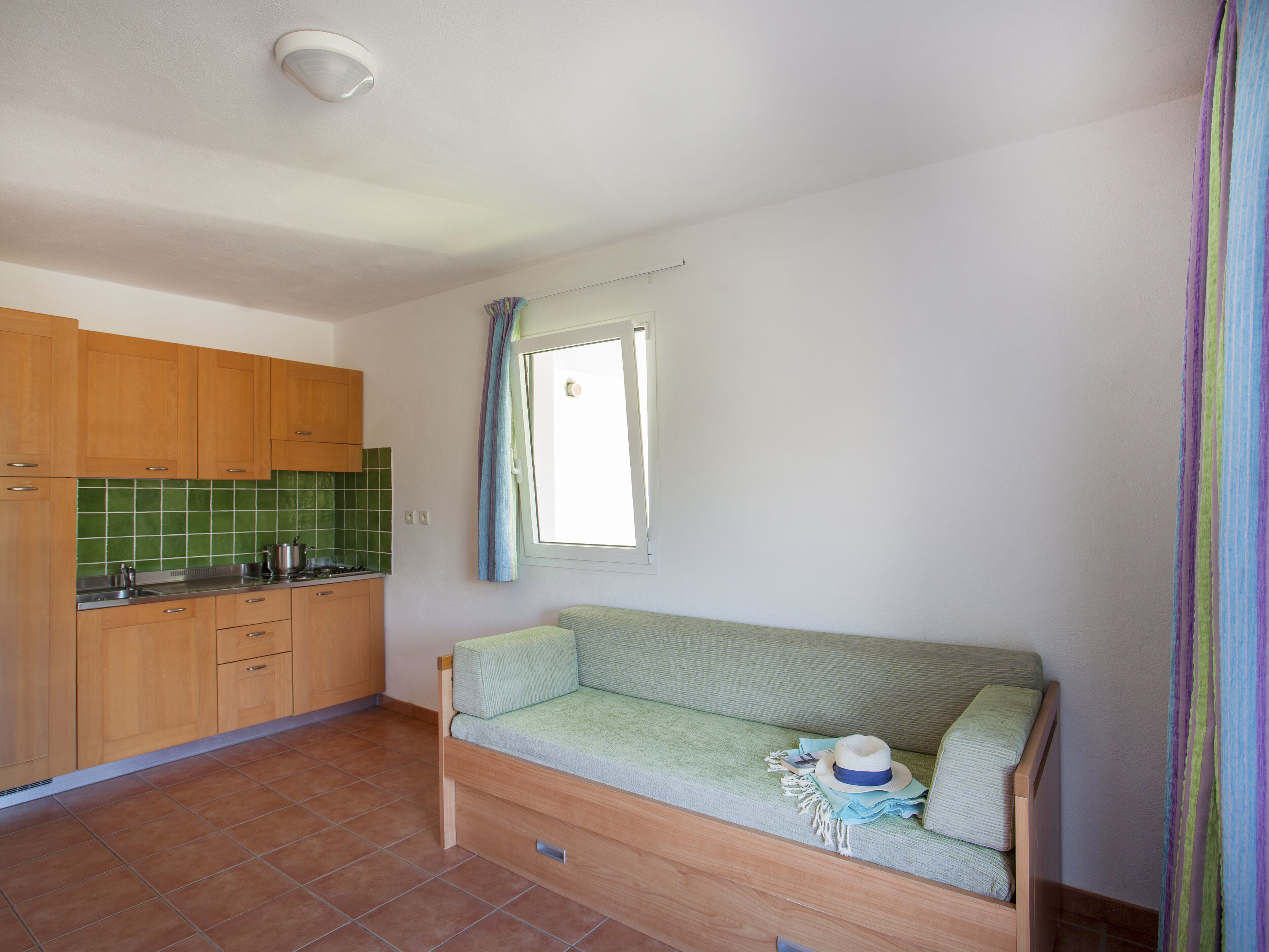 A compact kitchen with wooden cabinets, a green tiled backsplash, and a window with curtains.