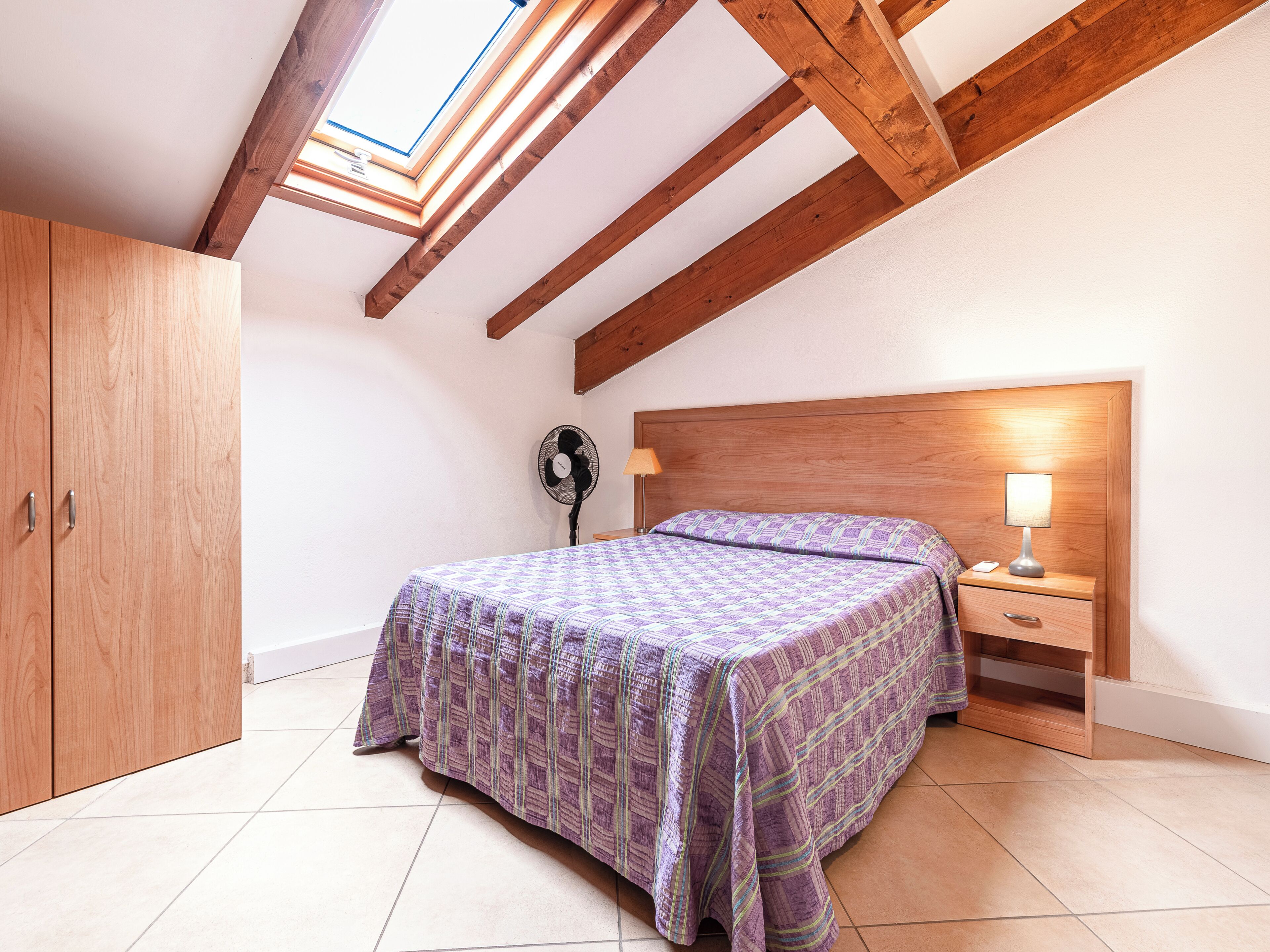 A bedroom with a bed, a nightstand, a lamp, a fan, and a wardrobe.