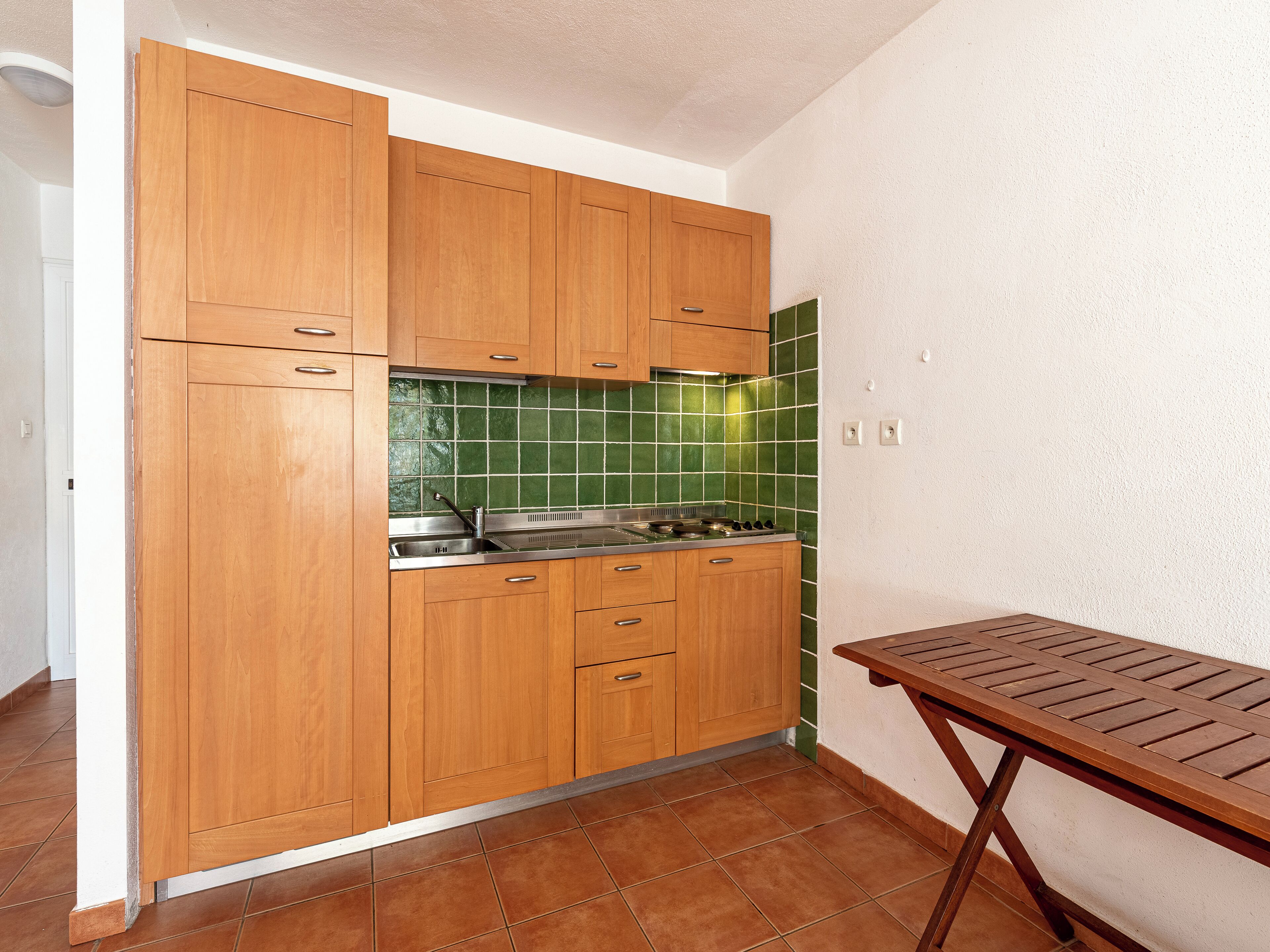A kitchen with wooden cabinets, a green tiled backsplash, a stainless steel sink, and a wooden table.
