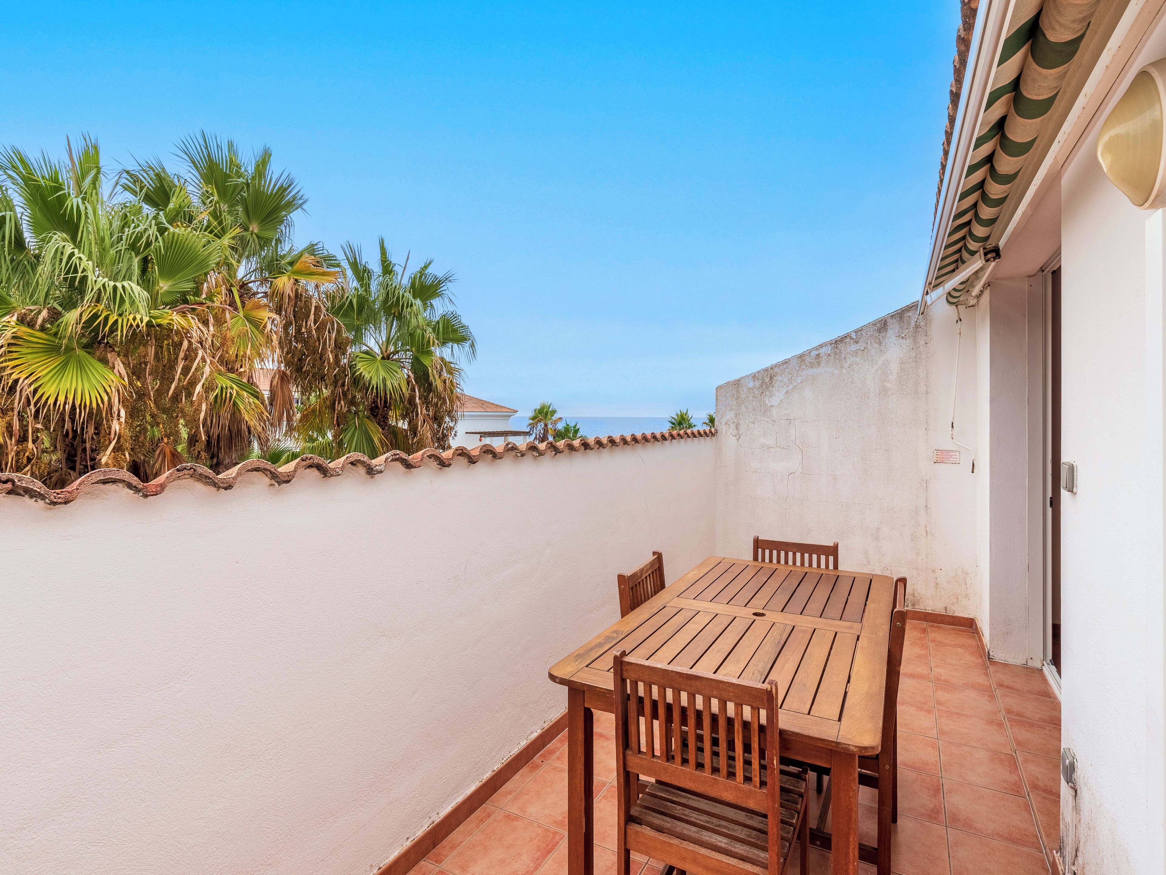 A patio with a wooden table and chairs, a white wall, and a view of the sea.