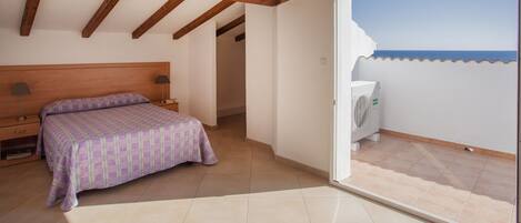 Un dormitorio con cama, mesita de noche, lámpara y vista al mar a través de una puerta de balcón abierta.