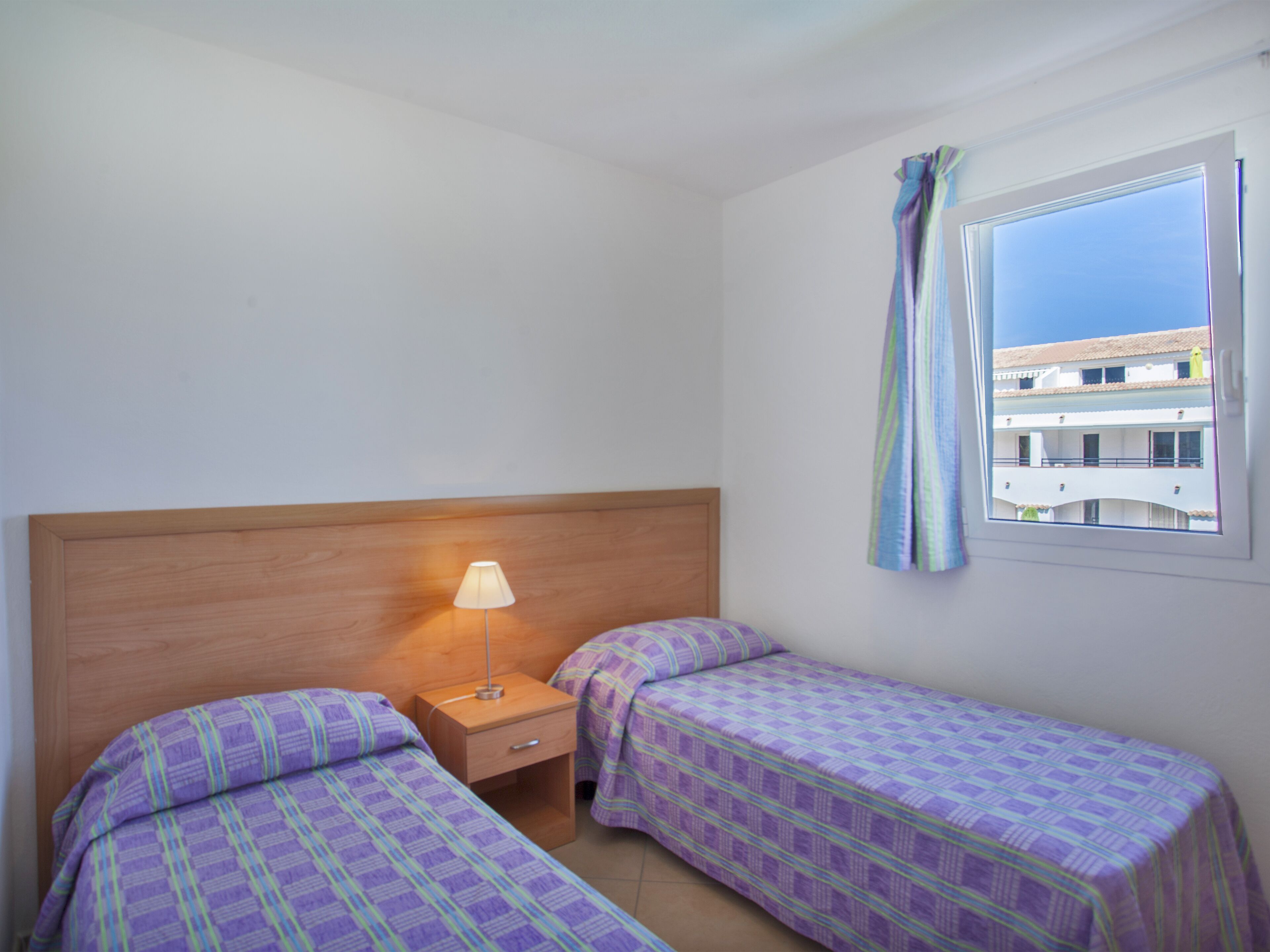 Een kleine, schone kamer met twee bedden, een nachtkastje, een lamp en een raam met uitzicht op gebouwen.