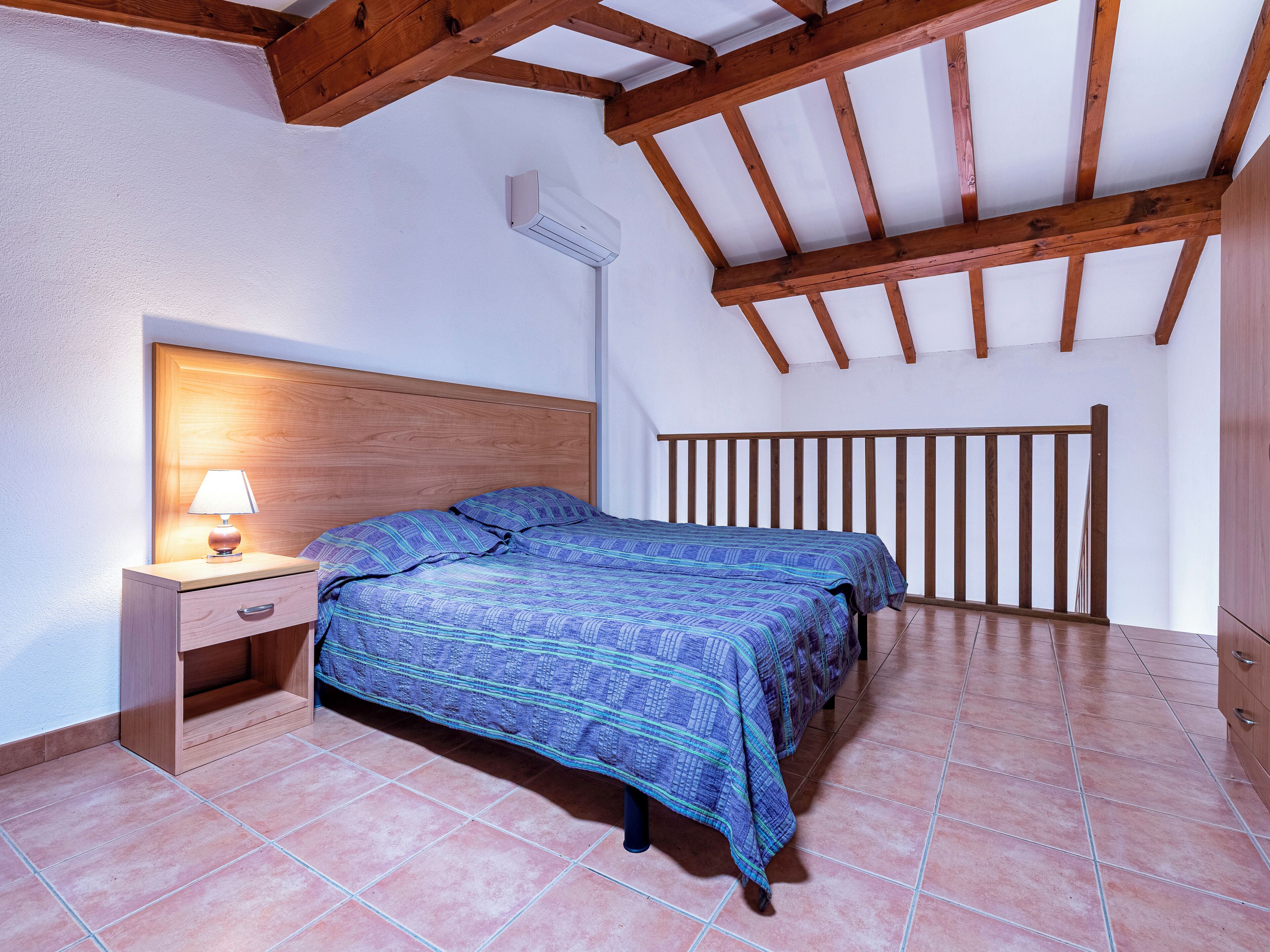 A bedroom with a wooden headboard, a bed with a patterned bedspread, a nightstand with a lamp, and a wooden ceiling beam structure.