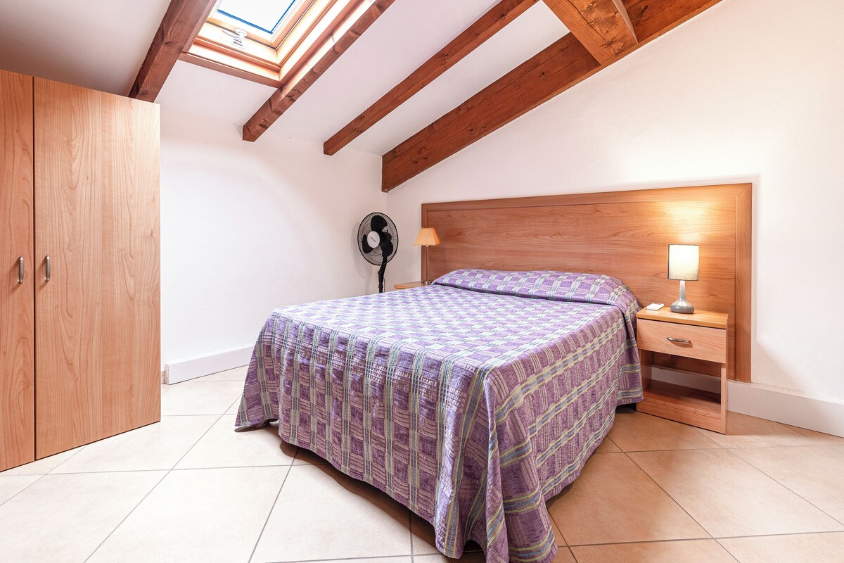 A bedroom with a sloped ceiling, wooden beams, a bed with a patterned bedspread, a bedside lamp, a fan, and a wardrobe.