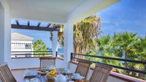 A wooden table set for breakfast with white plates, cups, and a fruit basket, overlooking a coastal view with palm trees and a building in the background. - Vacanceole Sognu Di Rena (San-Nicolao)