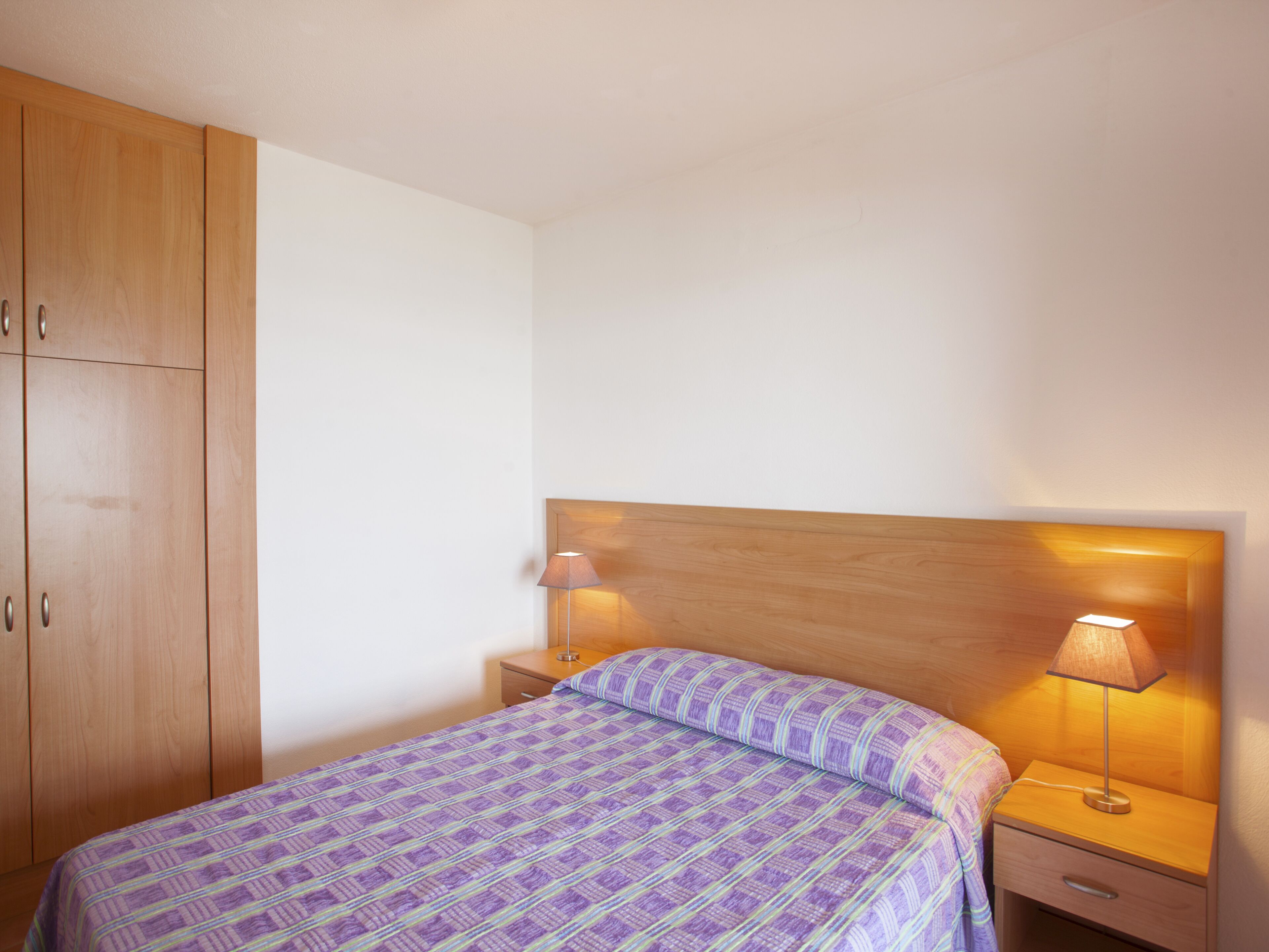 Una camera da letto con un'armadiatura in legno, un letto con copriletto a righe viola e blu, un comodino con una lampada e una testiera in legno.
