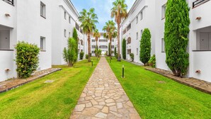 A courtyard with a stone pathway, surrounded by white buildings, palm trees, and neatly trimmed bushes. - Vacanceole Sognu Di Rena (San-Nicolao)