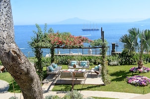 Un patio con mobiliario exterior, una pérgola con plantas en flor y vistas al mar, con un barco al fondo.