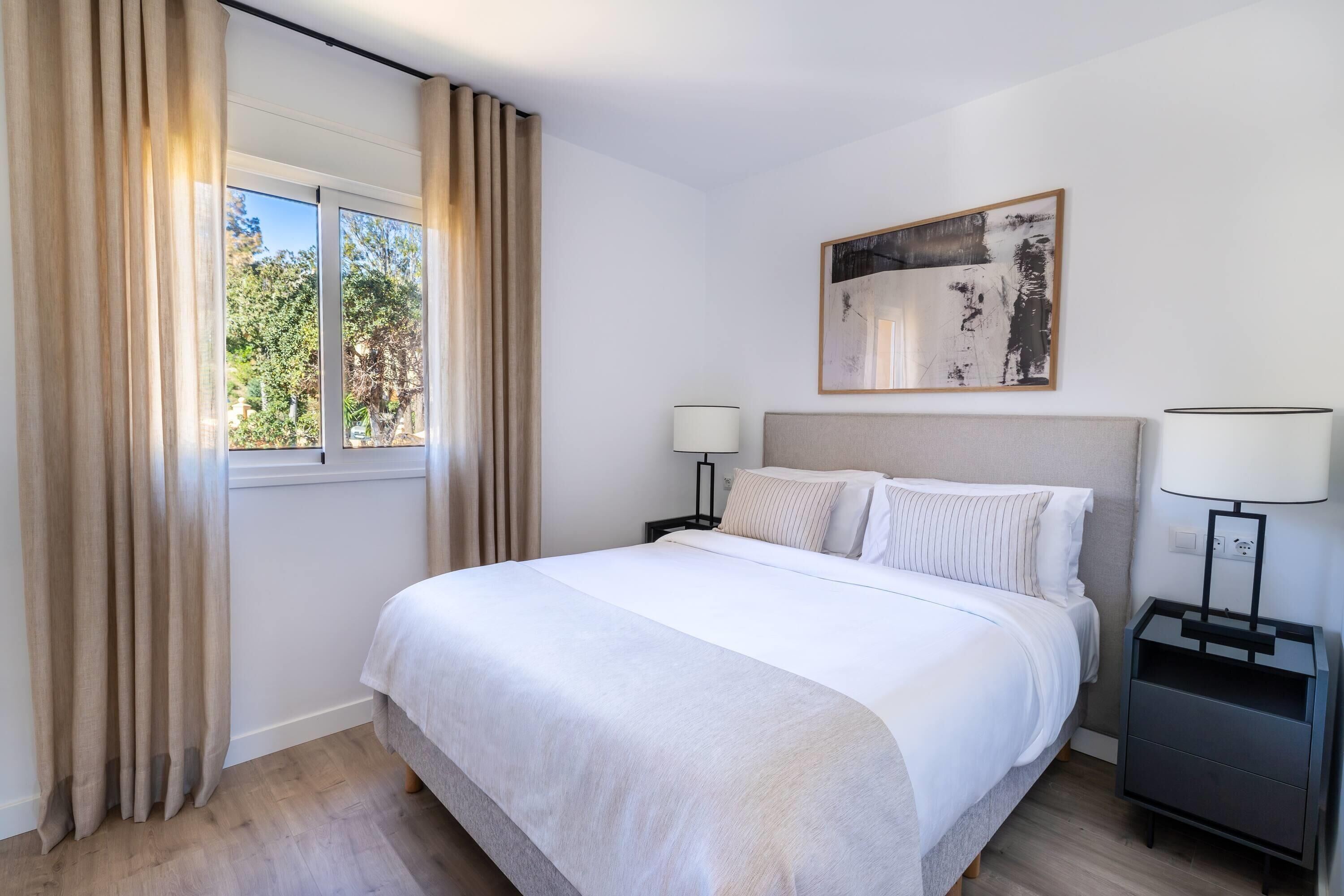 A hotel room with a bed, two bedside tables, a window with curtains, a wall-mounted artwork, and a view of trees outside.