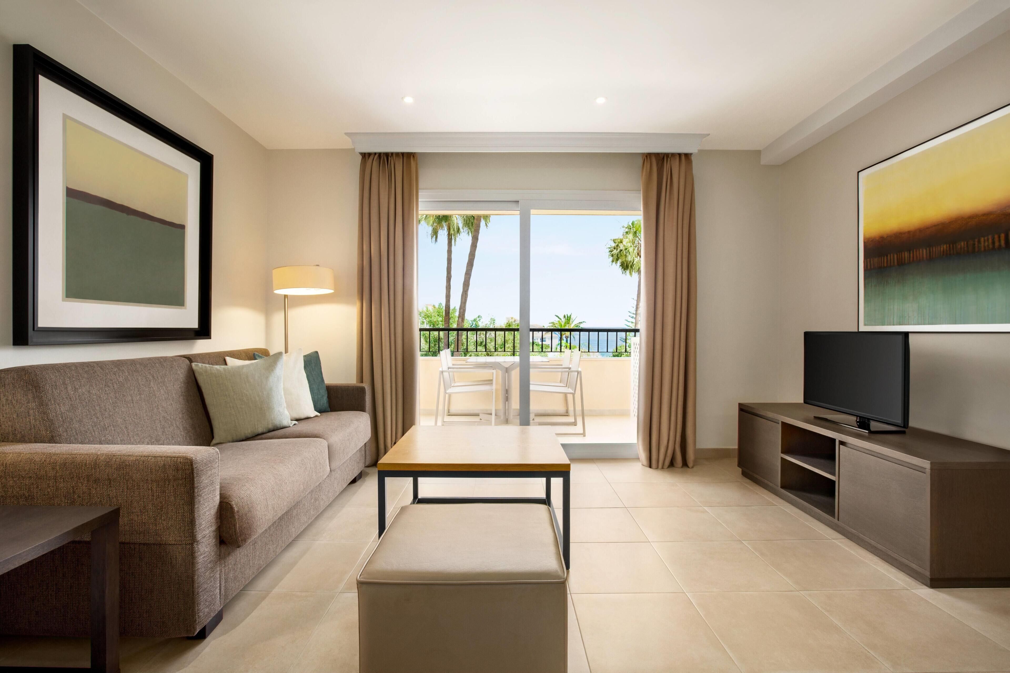A modern living room with a sofa, a coffee table, a TV on a stand, and a balcony with a view of palm trees.