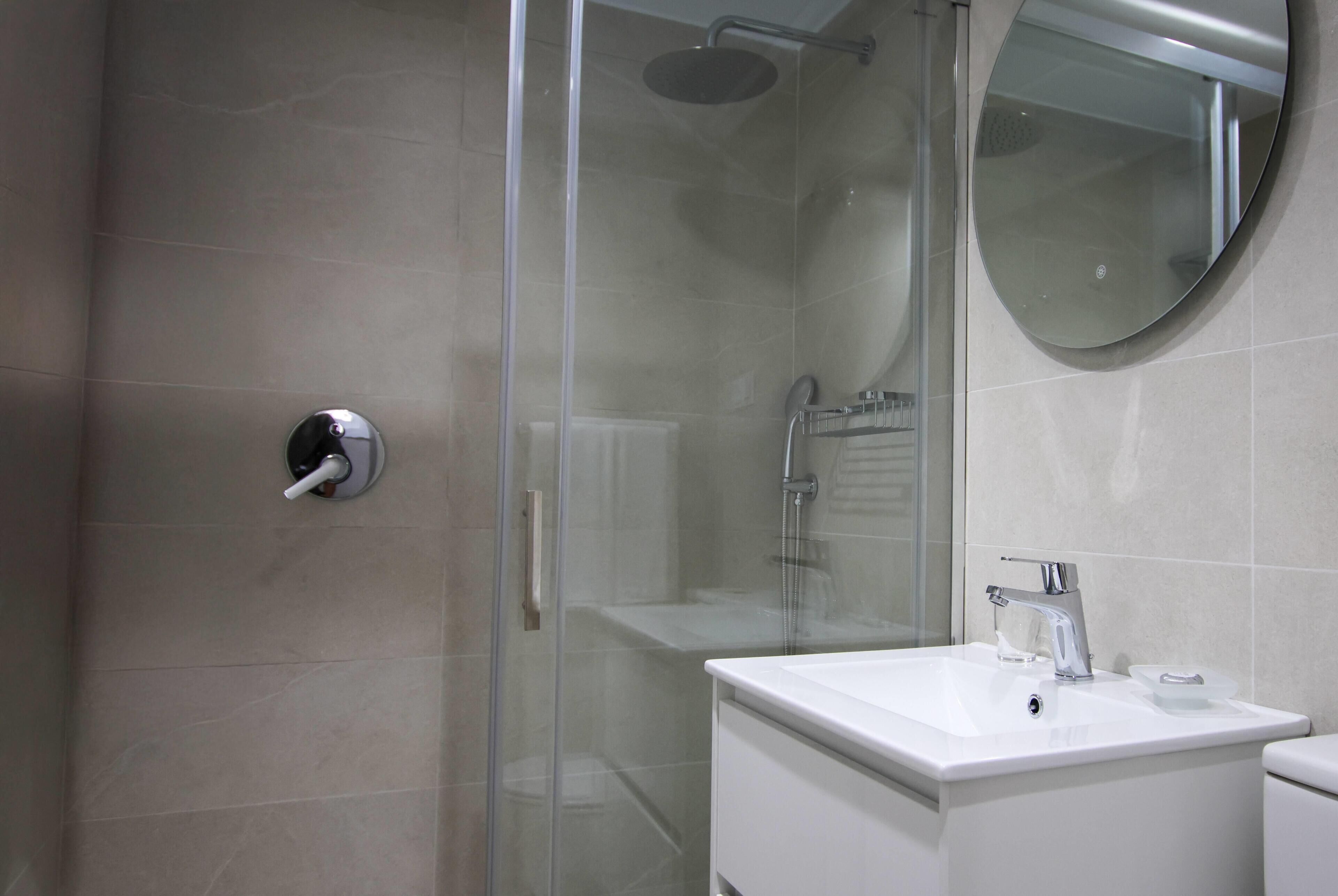 A modern bathroom with a glass-enclosed shower, a white sink with a chrome faucet, and a round mirror on the wall.