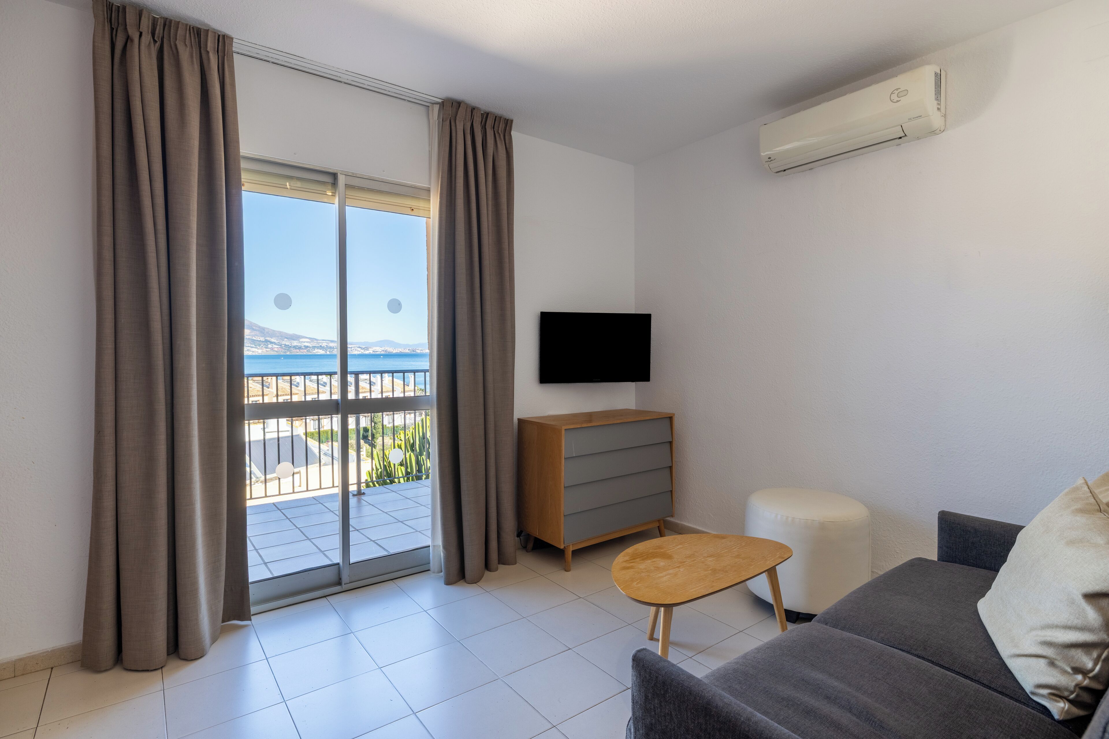 Un salon moderne avec un canapé gris, une table d’appoint en bois, un téléviseur fixé au mur et une porte-fenêtre coulissante donnant sur un balcon avec vue sur la mer.