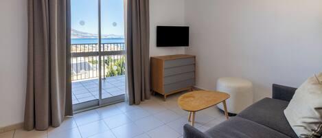 A modern living room with a grey sofa, a wooden side table, a TV mounted on the wall, and a sliding glass door leading to a balcony with a view of the sea.