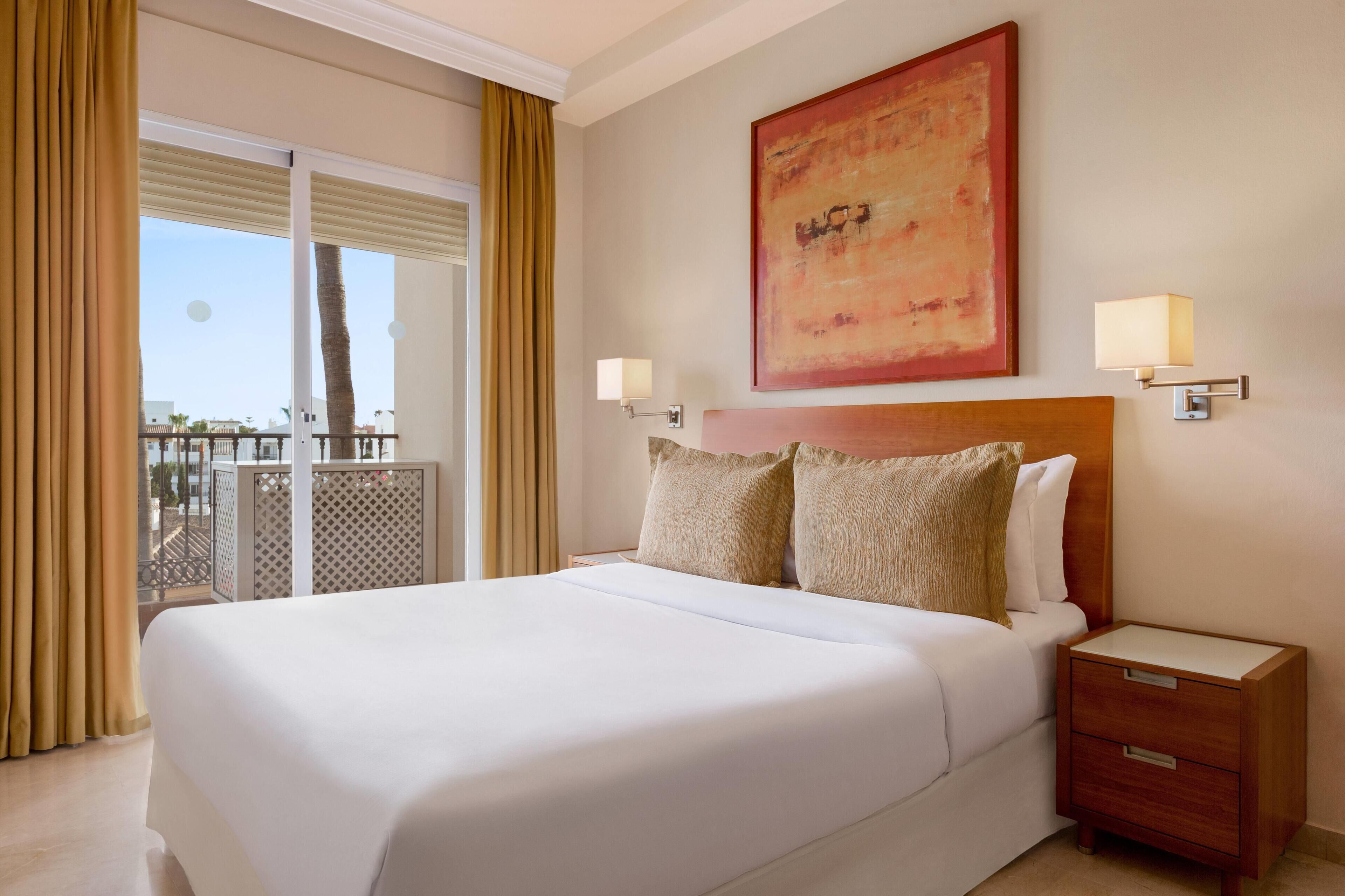 A hotel room with a large bed, a nightstand, a wall-mounted lamp, and a balcony view.