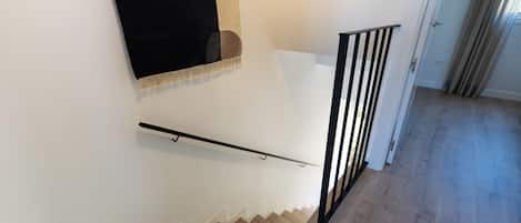 Una escalera con barandal negro, peldaños de madera y pared blanca.