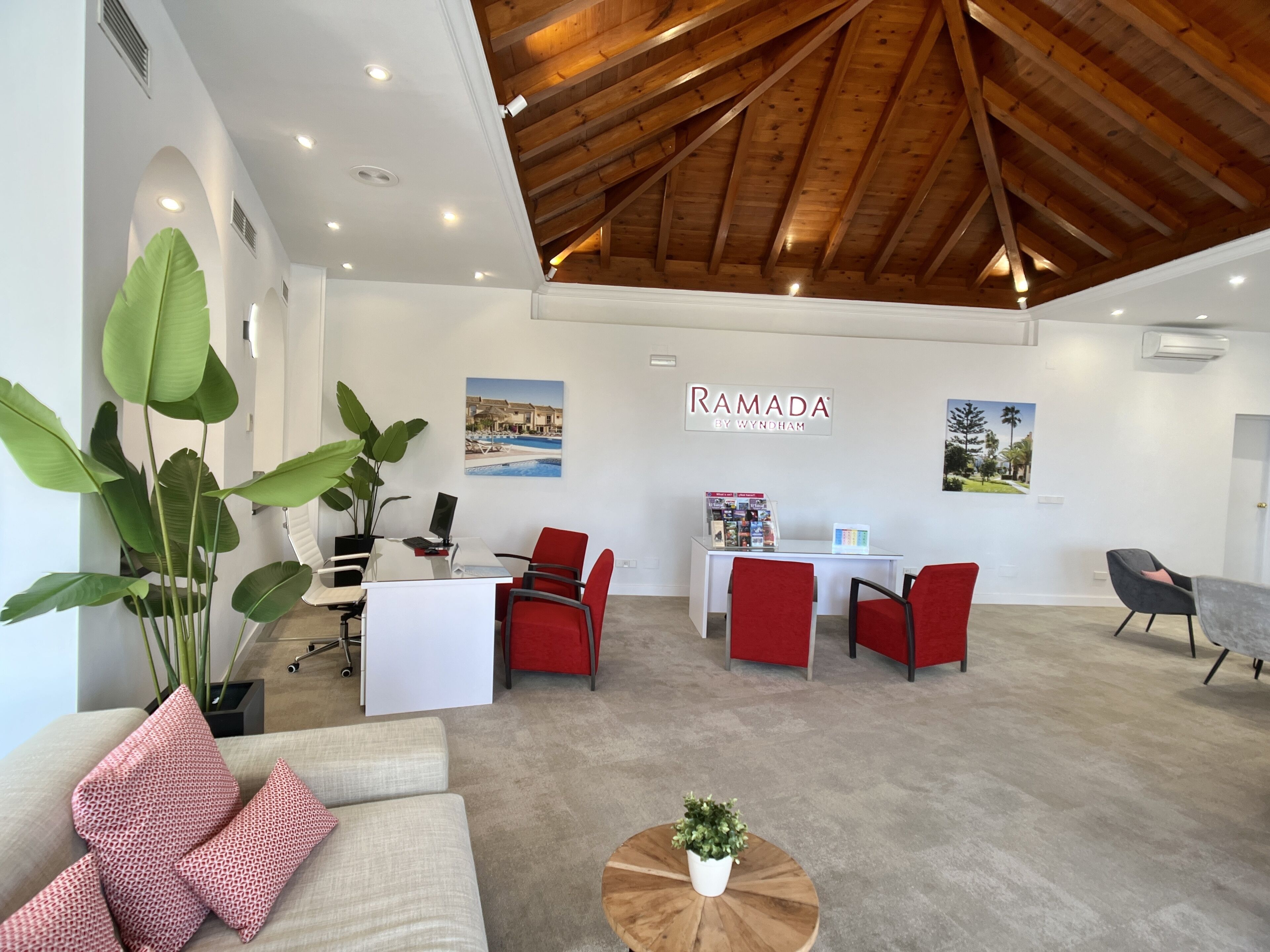 Un interior moderno con techo alto, una recepción, sillas rojas, un sillón gris y una planta grande en maceta.