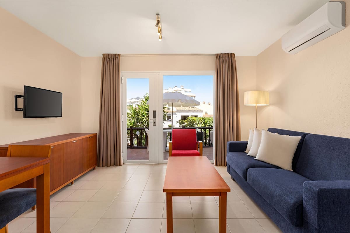 Habitació d'hotel amb taula de fusta, sofà blau, televisió i un balcó amb cadira vermella.