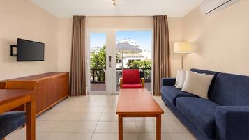 A hotel room with a wooden table, blue sofa, TV, and a balcony with a red chair.