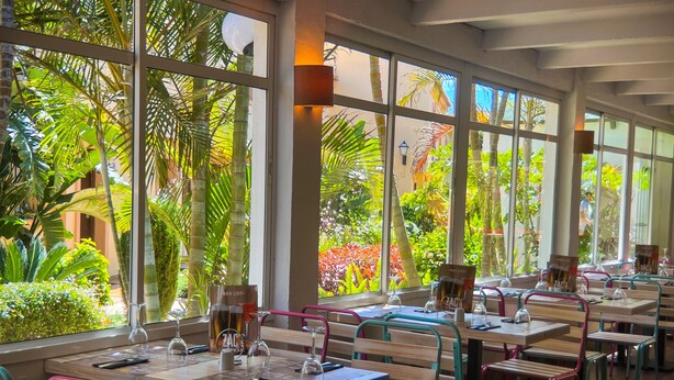 Un restaurante con amplios ventanales que ofrece vistas a exuberante follaje y palmeras. Las mesas están preparadas con copas de vino y menús.