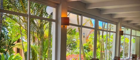 Un restaurante con amplios ventanales que ofrece vistas a exuberante follaje y palmeras. Las mesas están preparadas con copas de vino y menús.