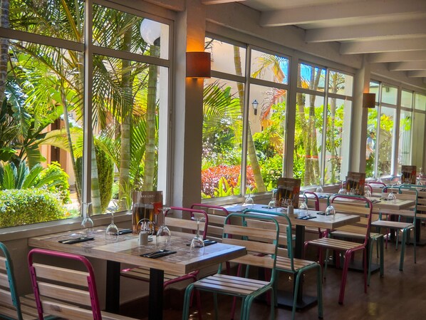 Un restaurante con amplios ventanales que ofrece vistas a exuberante follaje y palmeras. Las mesas están preparadas con copas de vino y menús.