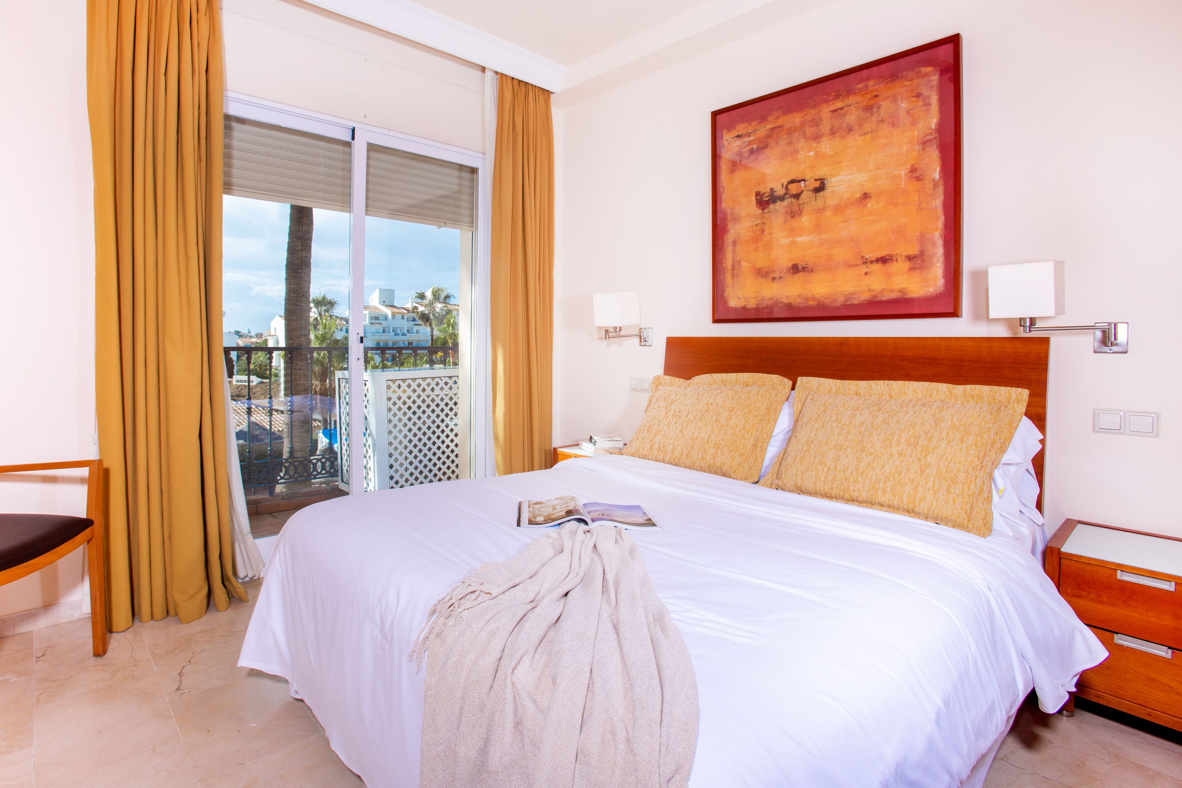 Un cuarto de hotel con una cama grande, un cabecero de madera, un balcón con cortinas amarillas y un cuadro en la pared.