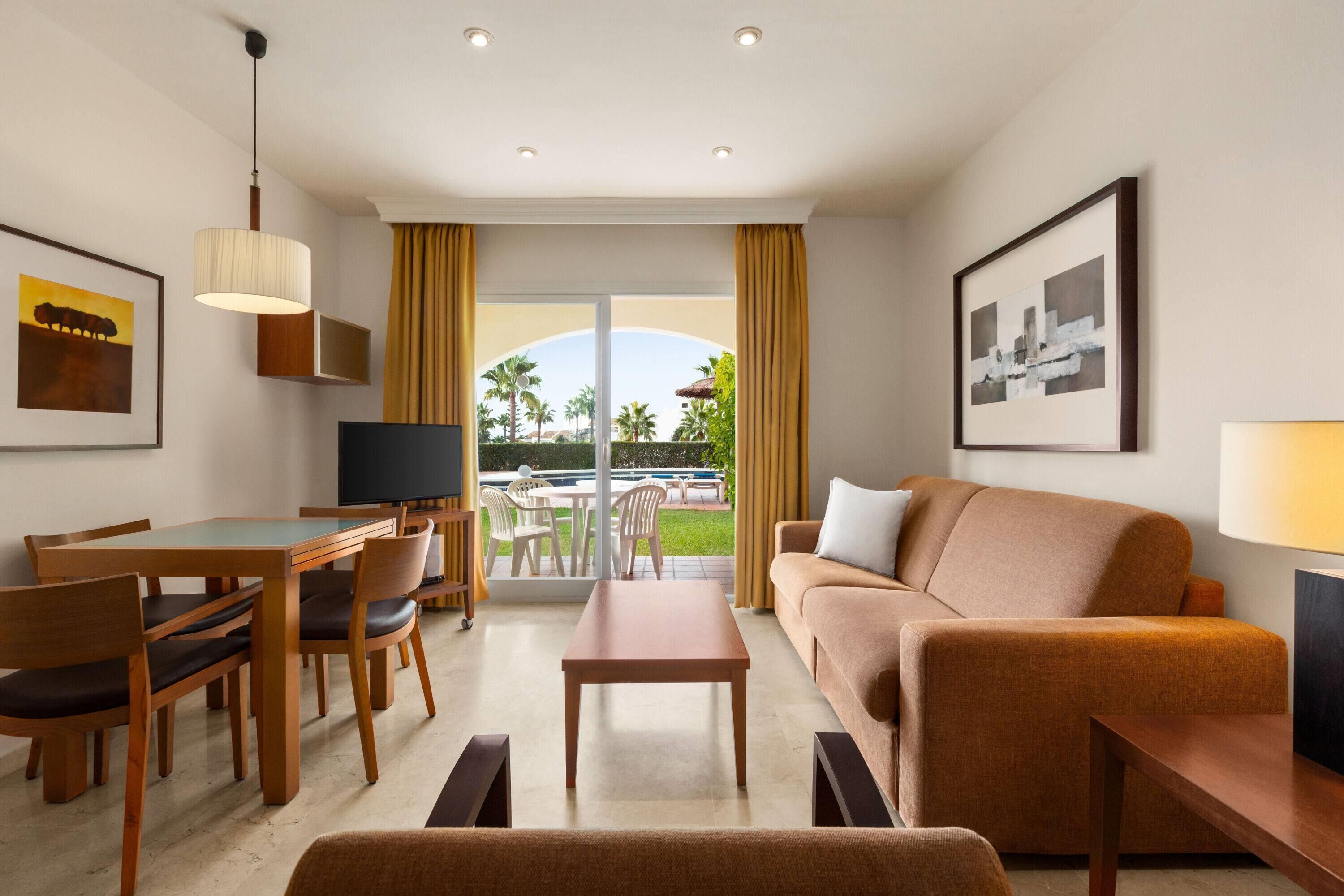 A hotel room with a dining area, a sofa, a TV, and a balcony with a view of palm trees.