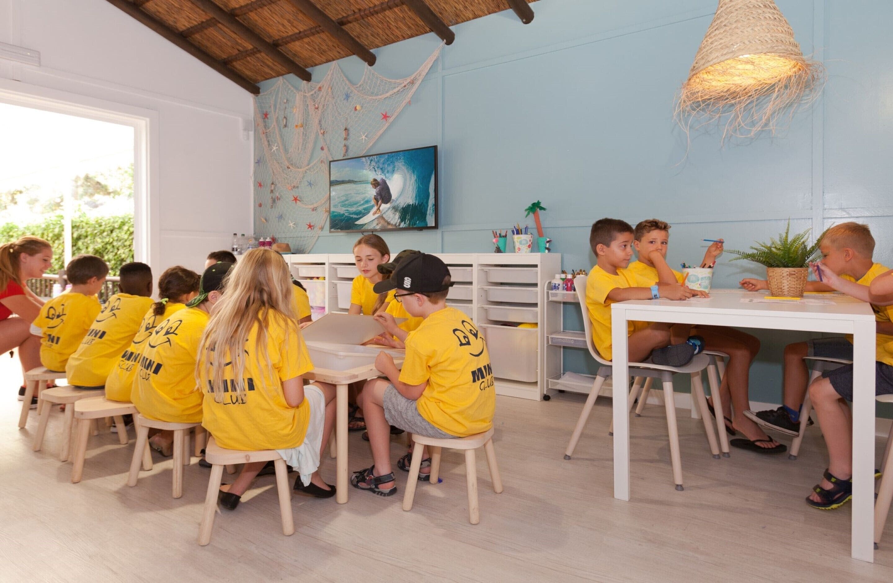A group of children in yellow shirts seated at a table in a room with a blue wall and a painting of a surfer.