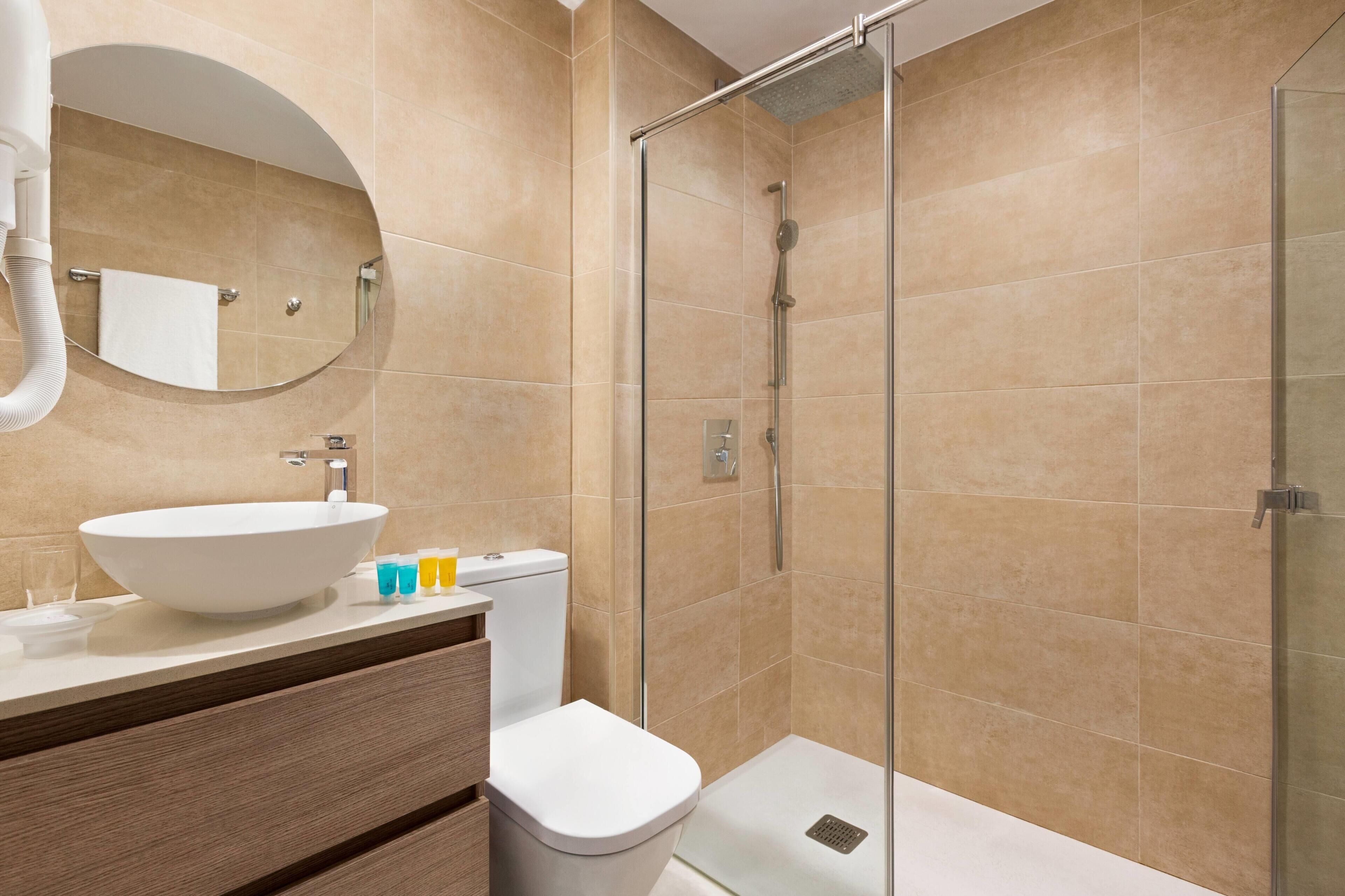 A modern bathroom with a round mirror, a white sink, a toilet, and a glass-enclosed shower.