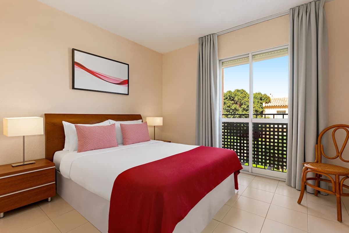 Habitació d'hotel amb llit, nits, una cadira, una finestra amb cortines i un balcó.