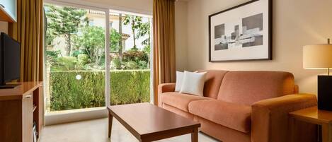 Une chambre d’hôtel avec un canapé, une table, une télévision, une lampe et une fenêtre donnant sur un paysage verdoyant.
