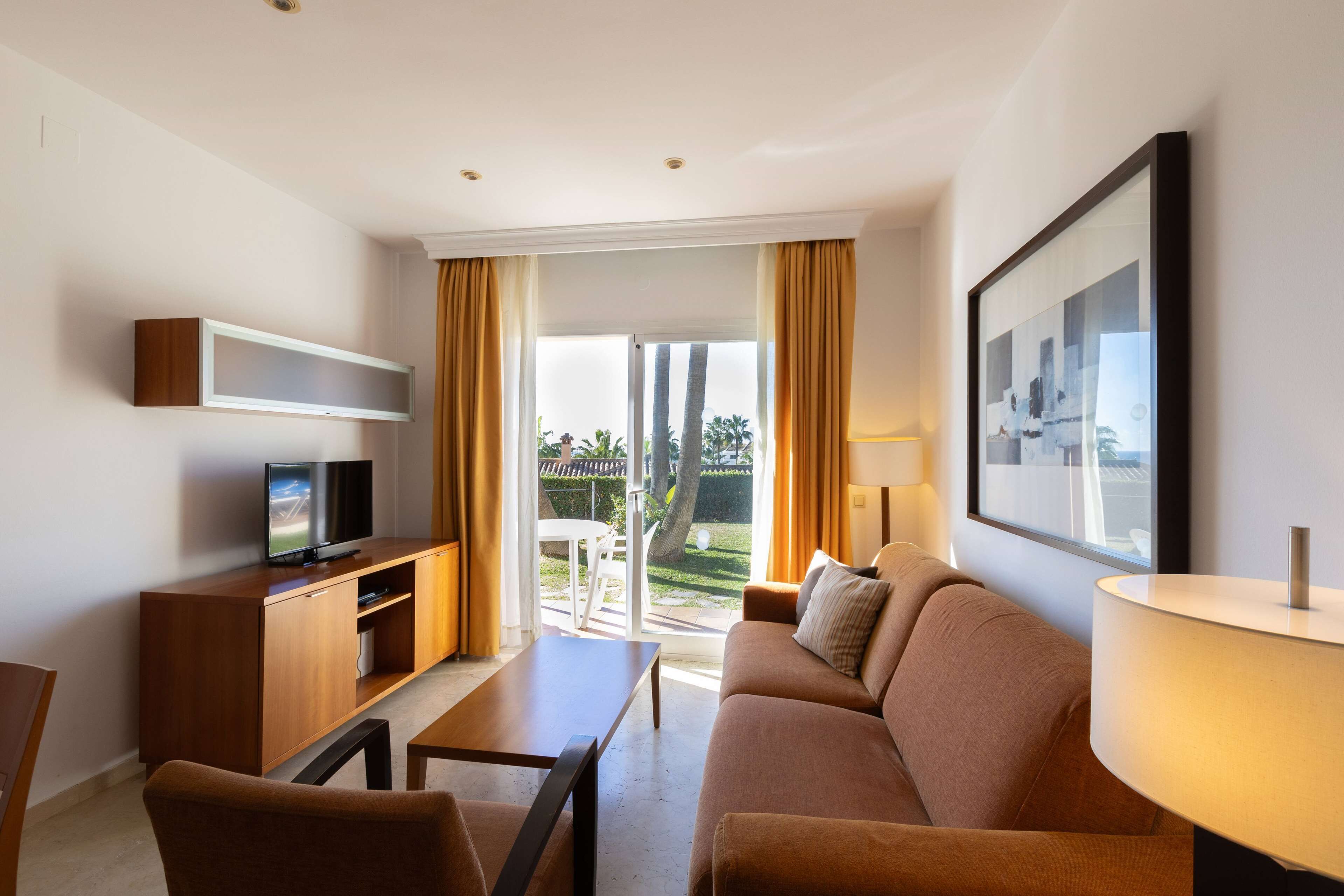 Habitación de hotel moderna con sofá, televisión sobre un mueble de madera, mesa y una ventana grande con cortinas.
