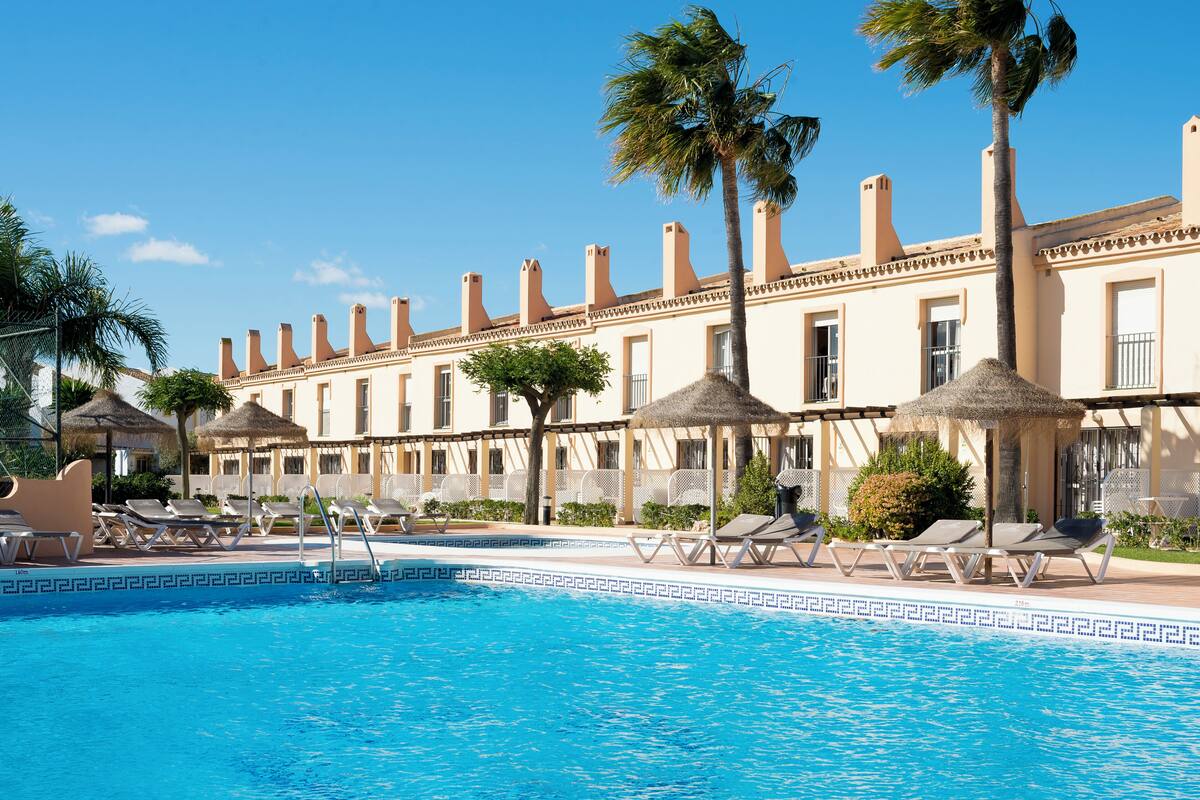 Un hotel amb piscina, gandules, palmeres i un cel blau clar.
