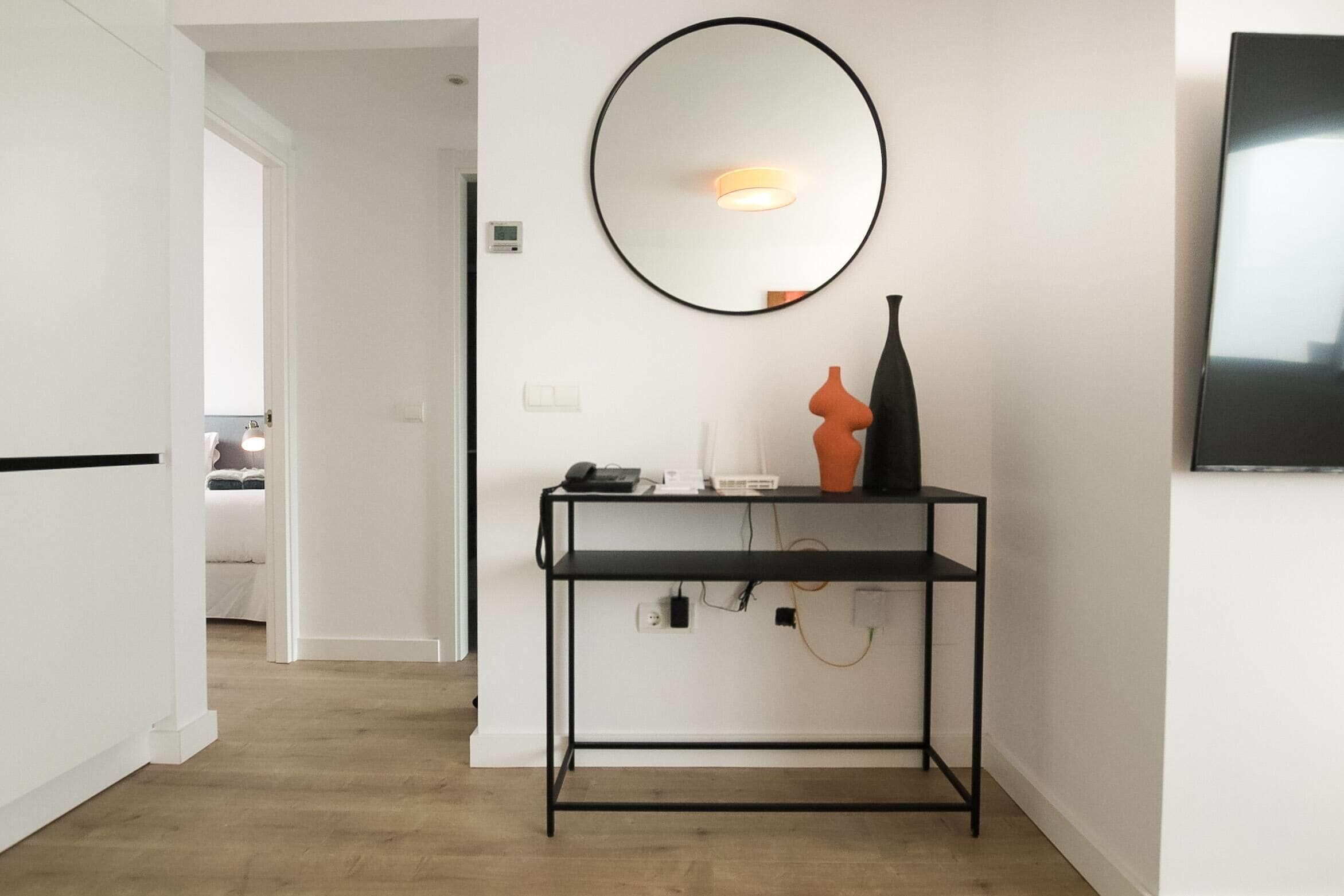 A modern room with a black metal console table, a round mirror, a flat-screen TV, and a door leading to another room.