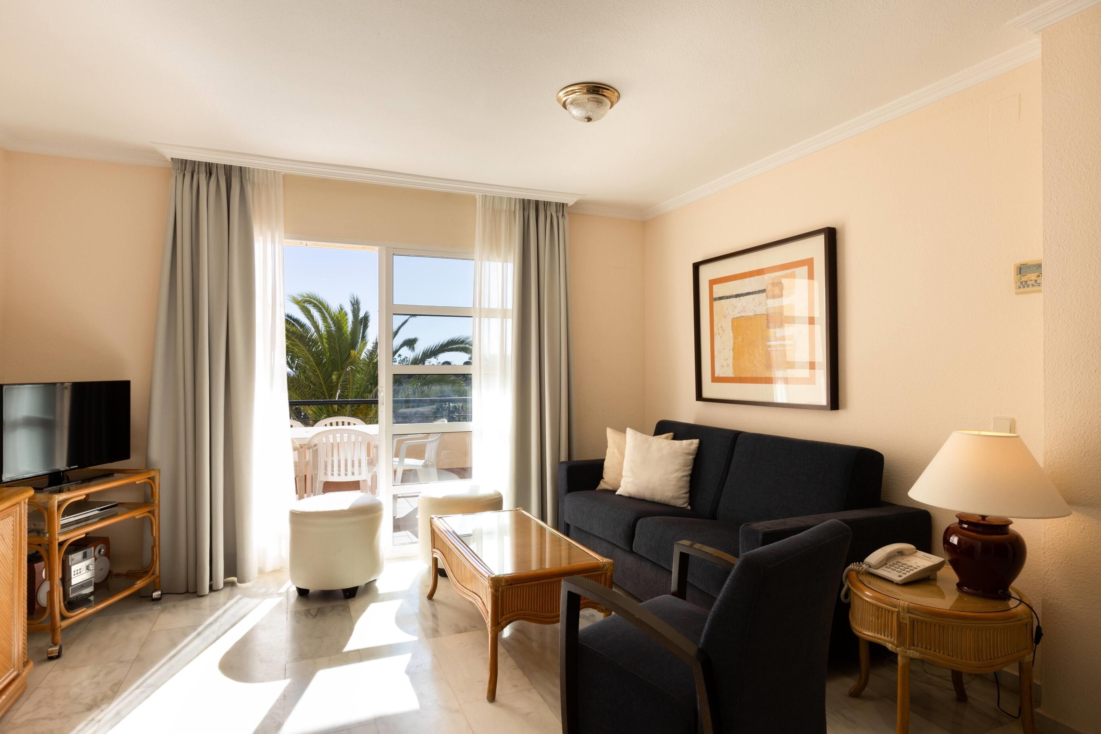 Habitación de hotel con sofá, televisor, lámpara, mesa, silla y balcón con vista a palmeras.