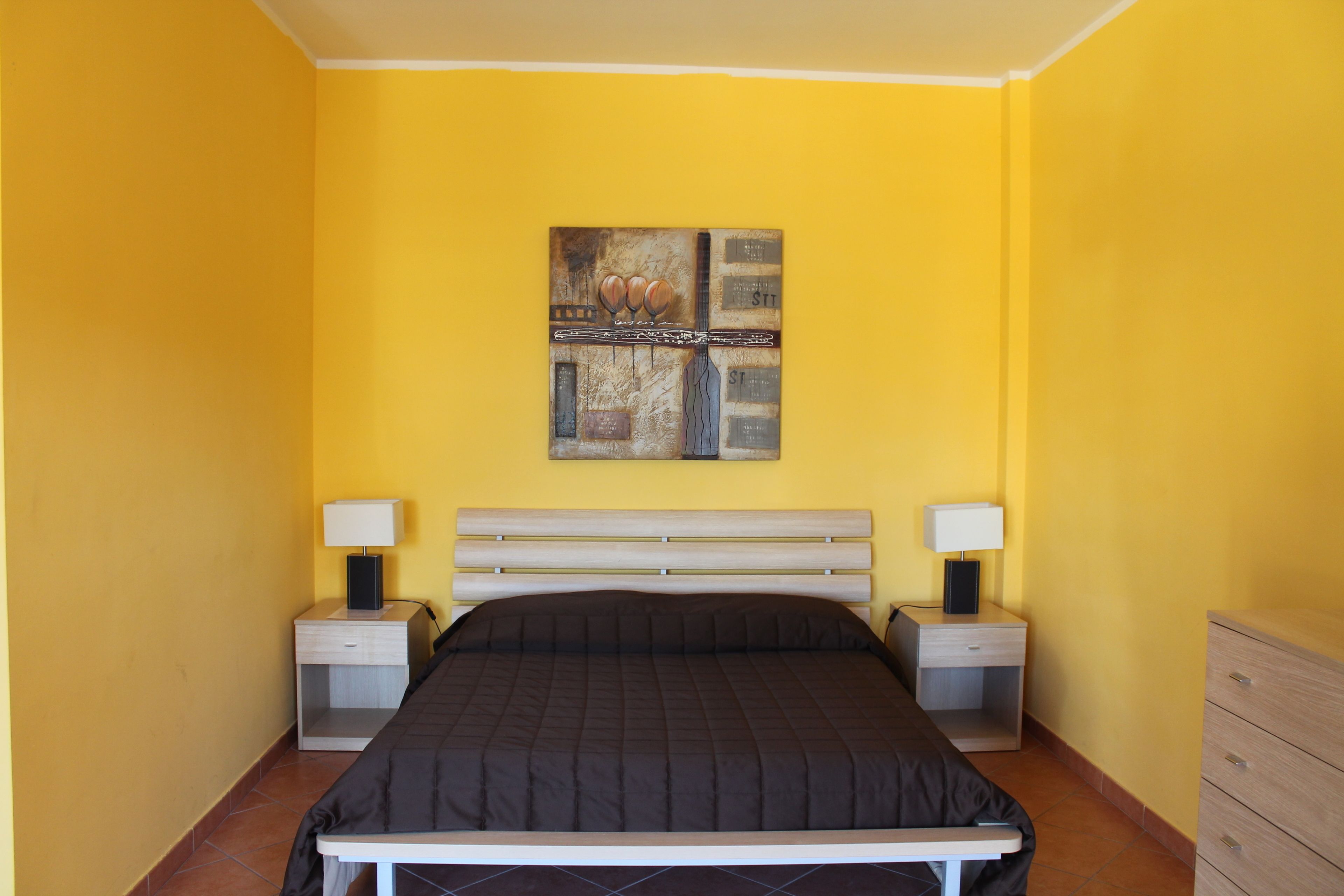 Ein Schlafzimmer mit gelben Wänden, einem Bett mit schwarzer Bettdecke, zwei Nachttischen mit Lampen und einer hölzernen Kommode.